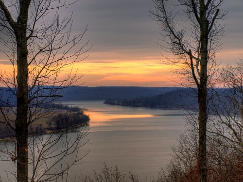 A serene river landscape at sunset, framed by bare trees and soft, muted colors in the sky.