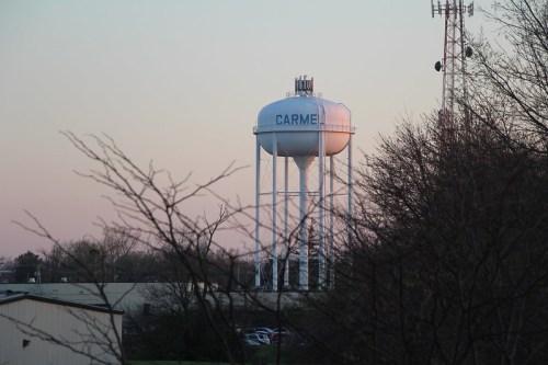 A water tower labeled "CARMEL" stands against a pastel sky, surrounded by trees and buildings.