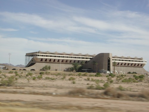 A large, modern building with a wavy roof, surrounded by desert landscape and sparse vegetation under a blue sky.