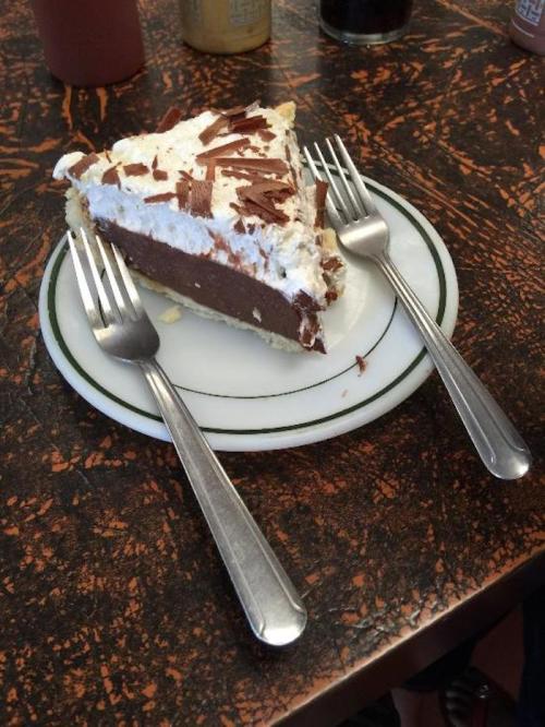 A slice of chocolate cream pie topped with whipped cream and chocolate shavings, served on a plate with two forks.