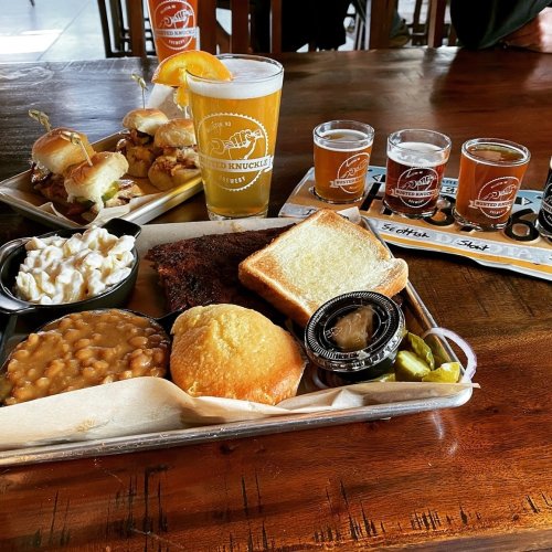 A tray of BBQ food with sliders, baked beans, cornbread, and a beer flight on a wooden table.