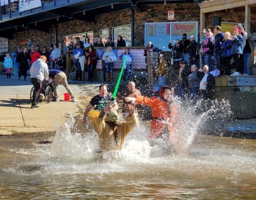 A group of people splashes into water during a fun event, with onlookers cheering from the shore.