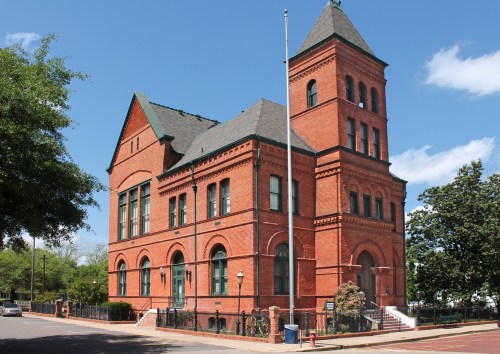 Historic red brick building with a tall tower, surrounded by trees and a clear blue sky.