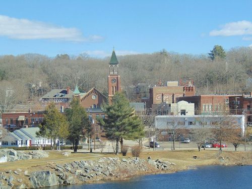 A scenic view of a small town with a clock tower, trees, and a lake under a clear blue sky.