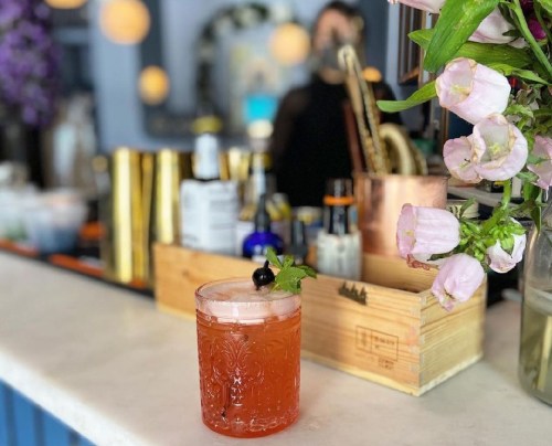 A vibrant cocktail garnished with a cherry and mint, placed on a bar counter with flowers and bottles in the background.