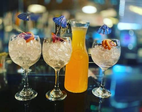 Three cocktails with ice and colorful umbrellas, alongside a pitcher of orange drink on a bar counter.