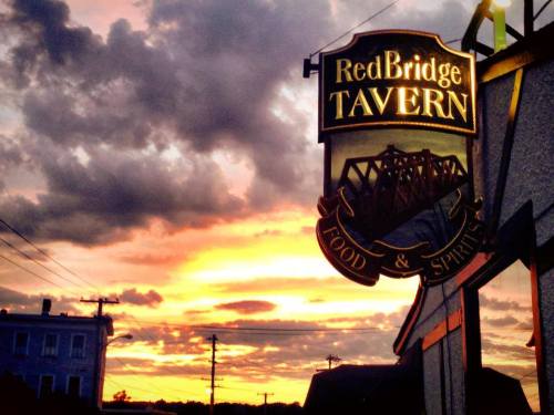 Sign for RedBridge Tavern against a vibrant sunset, with clouds and silhouettes of buildings in the background.