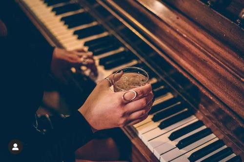 A hand holding a drink rests on a piano, with another hand playing the keys in a dimly lit setting.