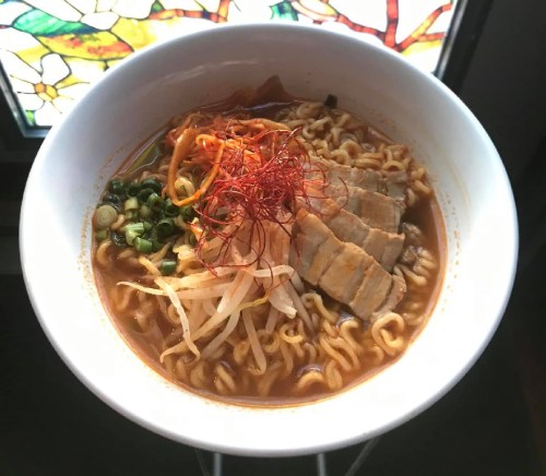 A bowl of ramen with sliced pork, green onions, bean sprouts, and red threads, set against a stained glass background.