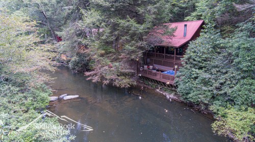 A serene river flows past cozy cabins nestled among lush trees, with a red-roofed porch overlooking the water.