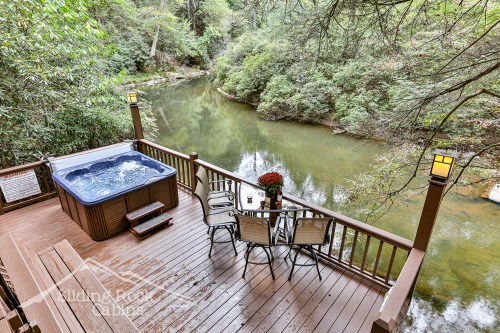 A serene deck overlooking a river, featuring a hot tub and a dining set with flowers, surrounded by lush greenery.