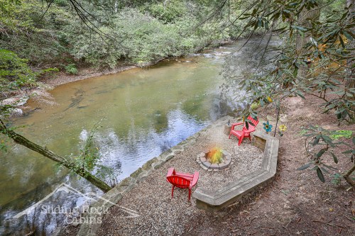 A serene riverside scene featuring a fire pit and red chairs surrounded by lush greenery.