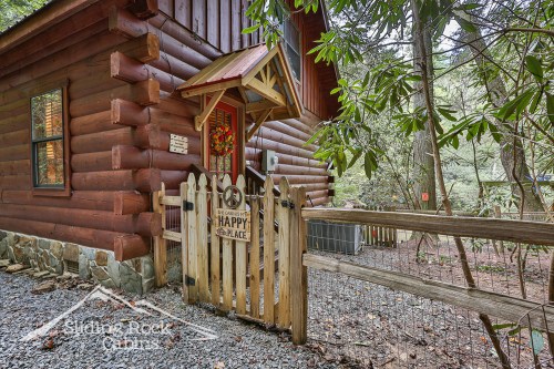 A cozy log cabin entrance with a wooden gate and a sign reading "Welcome to Our Happy Place," surrounded by trees.