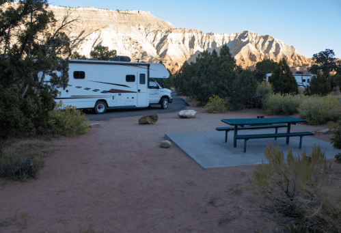 A campsite with an RV parked nearby, a picnic table on a concrete slab, and rocky terrain surrounded by trees and mountains.