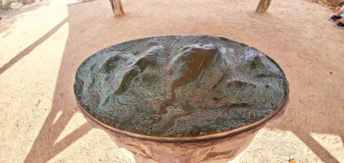 A circular bronze sculpture depicting a textured landscape, set on a stone pedestal in a sandy area.