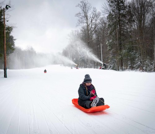 A person sleds down a snowy hill, surrounded by trees and snow-making machines in the background.
