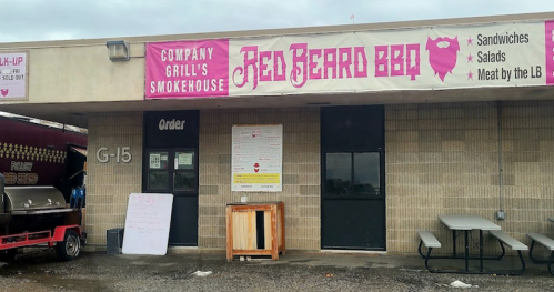 Exterior of Red Beard BBQ smokehouse with a sign displaying menu items and an outdoor seating area.