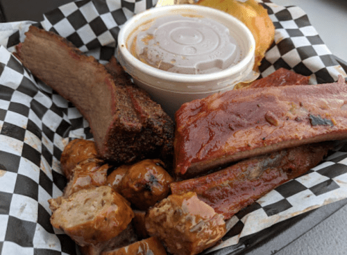 A basket of assorted barbecue meats, including brisket, ribs, and sausage, with a side of sauce and a slice of bread.