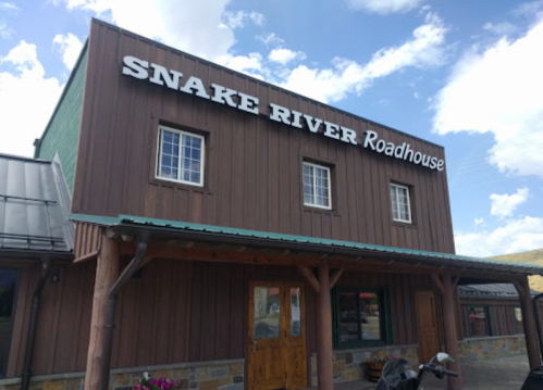 Exterior of the Snake River Roadhouse, a wooden building with a sign, under a blue sky with clouds.