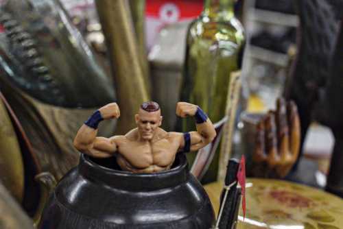 A muscular action figure flexes its arms while perched inside a vintage pot, surrounded by various objects.