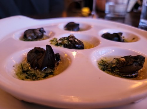 A white plate with six escargot, garnished with herbs and butter, served in individual compartments.