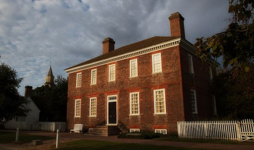 Historic brick house with white trim, surrounded by greenery and a picket fence, under a cloudy sky.