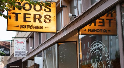 Sign for "Rooster's Kitchen" with a warm glow, located in a bustling street with nearby shops visible.