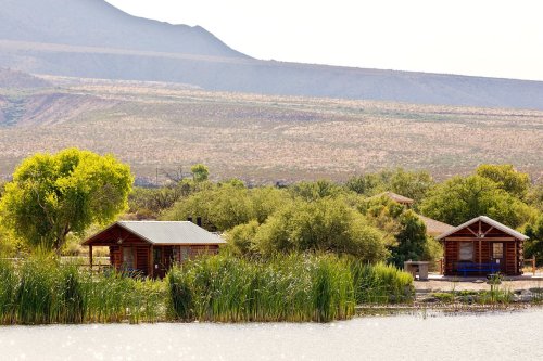 Two rustic cabins by a serene lake, surrounded by lush greenery and distant mountains under a bright sky.