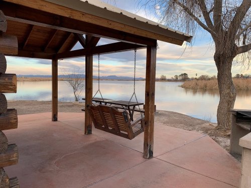 A wooden swing under a shelter by a calm lake, with trees and mountains in the background at sunset.