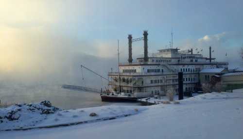 A snow-covered riverboat docked by a foggy river, with a cloudy sky in the background.