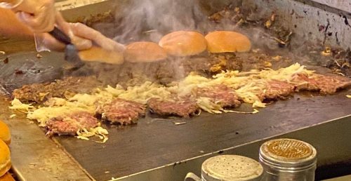 A grill with sizzling patties and buns, steam rising, as someone prepares a burger meal.