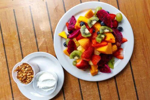 A colorful fruit salad with various fruits, served with yogurt and granola on the side, on a wooden table.