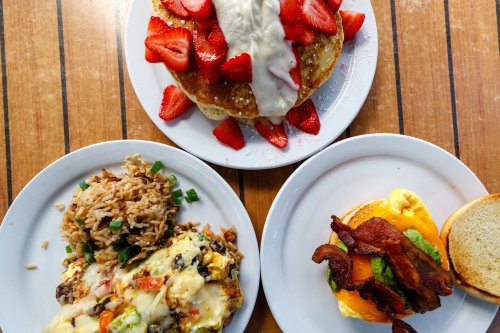Three plates of food: pancakes topped with strawberries and cream, a cheesy rice dish, and a bacon sandwich.
