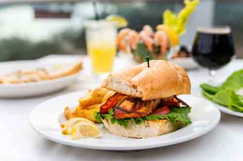 A delicious sandwich with grilled fish, lettuce, and lemon, served with chips on a white plate, with drinks in the background.