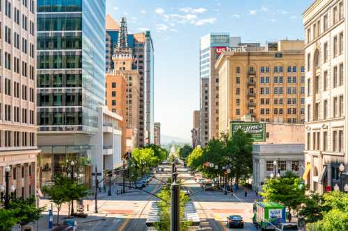 A vibrant city street lined with trees, tall buildings, and clear blue skies, showcasing urban life and architecture.