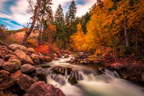 A serene river flows through a vibrant autumn forest, with colorful leaves and rocky banks under a cloudy sky.