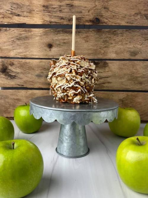 A caramel apple on a metal stand, surrounded by green apples, against a rustic wooden background.