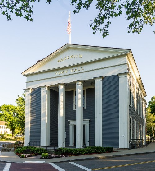 Historic Sandwich Town Hall features a classic design with columns and an American flag atop. Lush greenery surrounds it.