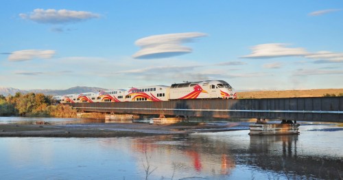 A train crosses a bridge over a river, with scenic clouds and mountains in the background.