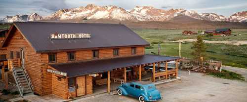 A rustic wooden hotel with a sign reading "Sawtooth Hotel," set against a backdrop of mountains and a blue vintage car.