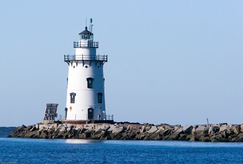 A white lighthouse stands on a rocky jetty, surrounded by calm blue waters under a clear sky.