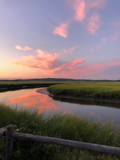 A serene landscape featuring a winding river, lush green grass, and a colorful sunset sky with pink and orange clouds.