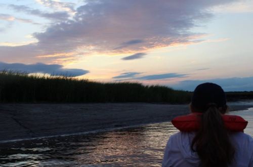 A person in a life jacket gazes at a colorful sunset over a calm waterway, surrounded by tall grass.