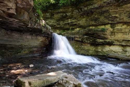 A serene waterfall cascading over rocky cliffs into a clear stream, surrounded by lush greenery.