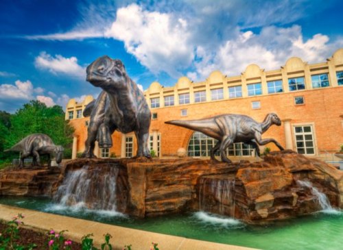 Three large dinosaur sculptures stand on a rocky base with water features, set against a blue sky and building backdrop.