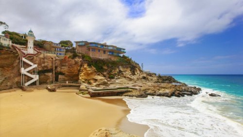 Scenic beach view with cliffs, a lighthouse, and modern homes under a partly cloudy sky. Waves gently lap the shore.