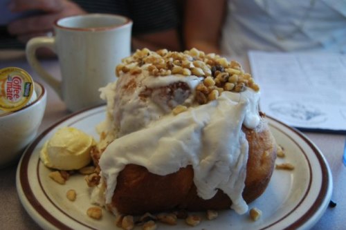 A large cinnamon roll topped with cream cheese frosting, chopped nuts, and a side of butter on a plate.