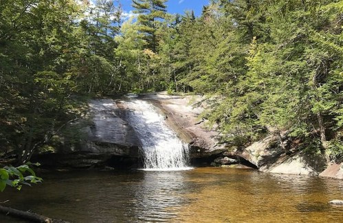 A serene waterfall cascades over rocks into a clear pool, surrounded by lush green trees and a blue sky.