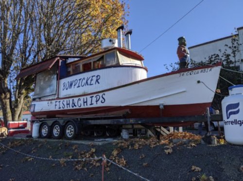A vintage boat named "Bowpicker" converted into a fish and chips restaurant, parked on a trailer with autumn leaves around.