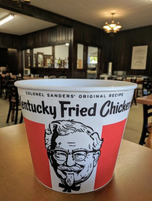 A bucket of Kentucky Fried Chicken with Colonel Sanders' image, set in a rustic dining area.
